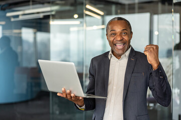 Fototapeta premium Mature businessman smiling and raising a fist in triumph while holding a laptop in a modern office, celebrating professional success, achievement and confident accomplishment