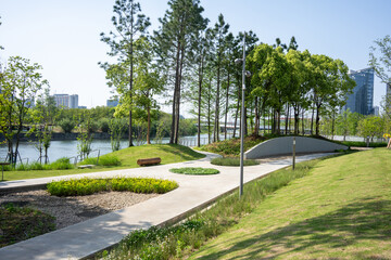 Fototapeta premium Winding pathway through green park lawn with trees and lake near city buildings