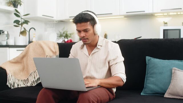 Indian man at home edits short video on laptop with wireless headphones trims clips adds cut and saves. Arabian guy on sofa works freelance checks timeline exports file and smiles with approval proud
