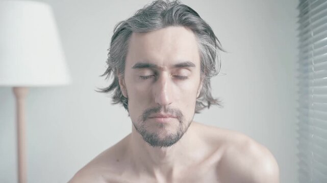 Serious look of a beautiful young man in a softly lit minimalistic bedroom, displaying vulnerability and thoughtfulness through introspective expressions and reflective gazes