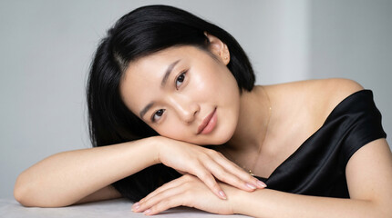 Obraz premium portrait of a young woman. Soft beauty portrait of a young Asian woman with short black bob haircut and natural makeup, resting her head gently on crossed arms, elegant black off-shoulder satin outfit