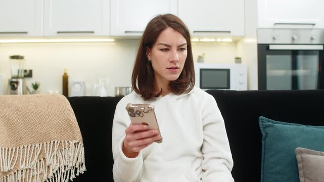 Young woman at home reads negative phone alert frowns whispers why did this happen feels shock and fear. Caucasian girl on sofa looks down worried holds smartphone tight reads message again seeks help