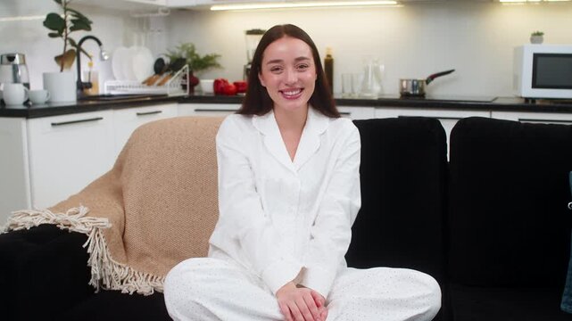 Young woman at home waves to camera smiles and greets with hello raises hand friendly vibe. Caucasian girl on sofa sits relaxed looks to lens happy mood kind gesture welcome sign positive energy yay.
