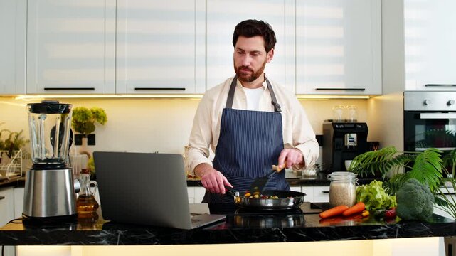 Middle-aged man cooks vegetables on frying pan seasoning food while watching recipe video on laptop in home kitchen. Young guy smiles feeling proud about healthy diet cooking for fitness weight loss.