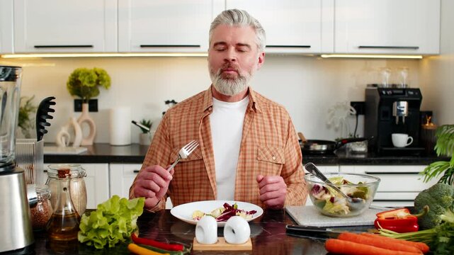 Middle-aged man tasting fresh homemade vegetable salad smiling after healthy meal prep in home kitchen. Mature guy enjoys diet food and mindful eating for weight loss health fitness and heart health.