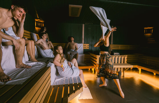 Female sauna master waving a towel during an aufguss ritual for a group of young people in a dry wooden sauna