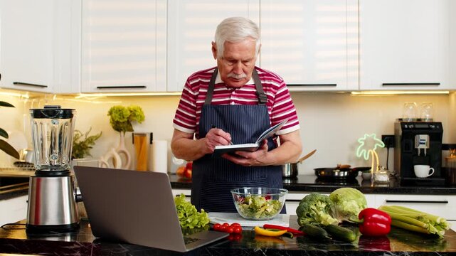 Senior old man prepares vegetable salad watching laptop recipe and writing notes in notebook for diet goals. Elderly grandfather tracks calories and nutrition in kitchen feeling focused and motivated.