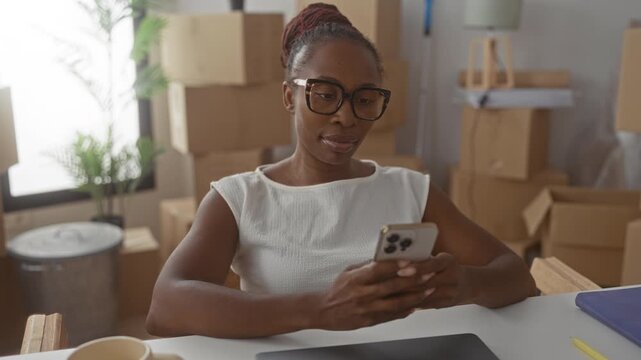 Young woman wearing glasses sits at desk holding smartphone among stacked moving boxes in building with potted plant and notebook visible; fresh start contentment.