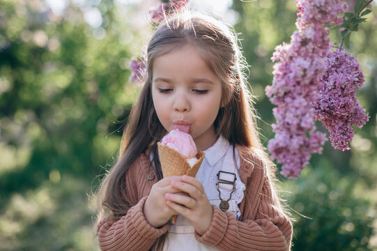 Cute little girl eating pink ice cream cone near blooming lilac flowers in sunny spring garden outdoors