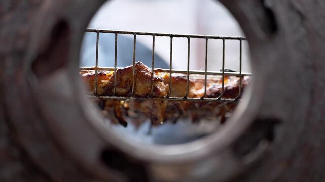 Chicken skewers sizzle inside a metal oven, viewed through a circular opening with sharp focus on the charred meat texture. Concept of culinary preparation and artisanal cooking methods.