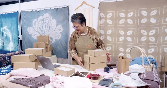 Senior asian businesswoman carries stacked parcel boxes, smiles, moves shipment across home studio, checks online order status on laptop, prepares delivery pickup, keeps invoice and receipt ready.