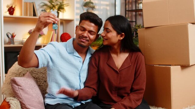 Indian couple man and woman sitting on home sofa show keys and smile with joy. Young family man and woman sitting on home sofa celebrate moving housing rental or purchase with boxes and fresh plans.