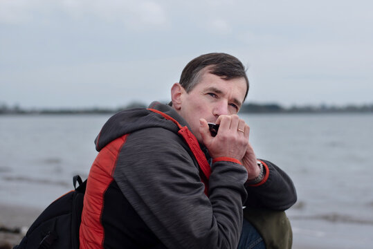 A contemplative man plays a harmonica while sitting by waterfront, evoking solitude, reflection and quiet music on an overcast day near a calm river, playing the harmonica