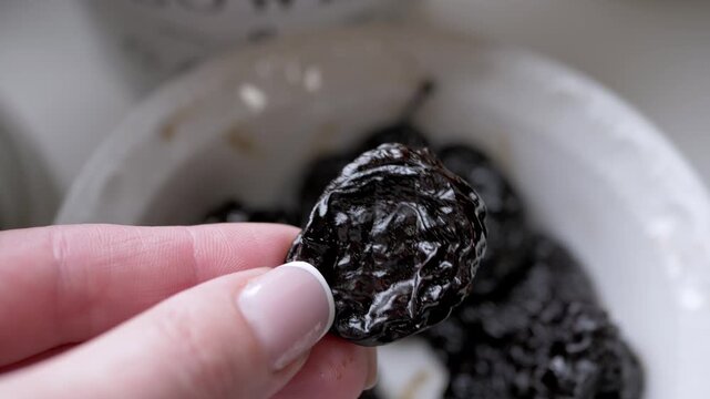A manicured hand holds a single dark dried prune plum over a blurry white bowl filled with similar fruit. Concept of healthy snacks, fiber-rich nutrition, organic food, and natural