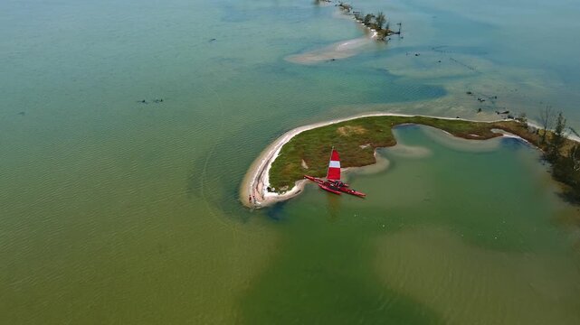 High-angle anti-clockwise drone orbit around a red and white trimaran sailboat moored on a curved sandbank surrounded by the shallow green waters of Araruama Lagoon in Rio de Janeiro, Brazil.