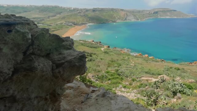 A rocky foreground frames of Mixta Cave in Gozo frames the vibrant turquoise sea and sandy Ramla beach below. Ideal for travel promotions or nature documentaries.