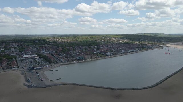 A smooth drone shot glides over West Kirby Marine Lake, capturing sailboats drifting across calm water beneath a bright blue sky dotted with soft clouds.