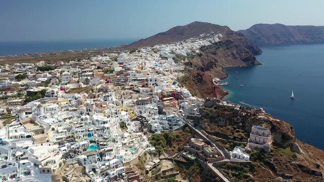 Beautiful wide shot of Oia, Santorini, featuring iconic white houses, blue-domed churches, and dramatic red volcanic cliffs slanting down into the deep blue Mediterranean Sea on a clear day.