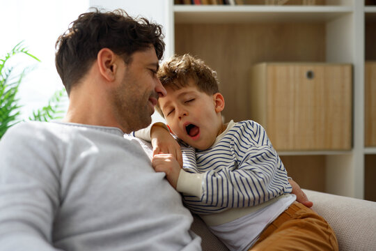 Sleepy little boy yawning while cuddling with father on sofa