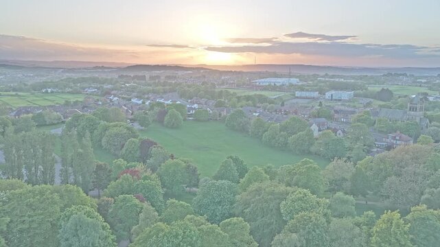 Locke Park and Barnsley stretch across South Yorkshire, Yorkshire and the Humber, England, drone pullback aerial shot above mature woodland, residential districts, and glowing sunset horizon.