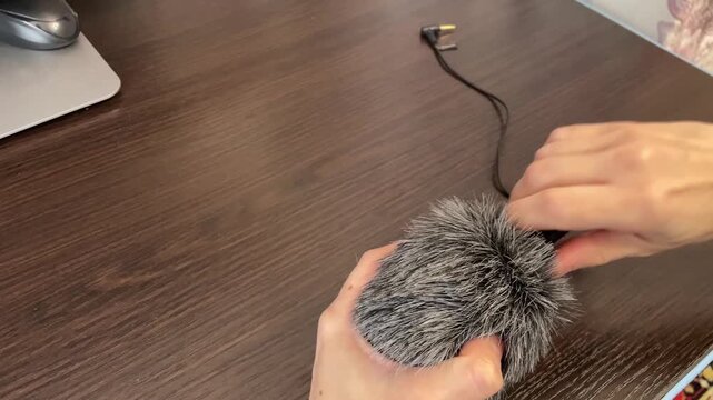 Furry windscreen on tabletop microphone caucasian hands secure foam connector to mic, usb cable visible, studio prep for podcast or vlog recording, textured gray fur windjammer, focused closeup