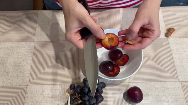 Hands slicing ripe plums into bowl, preparing fresh fruit snack on patterned tablecloth, grapes placed nearby, paring knife used to halve plums and remove pits, casual home kitchen setting with soft