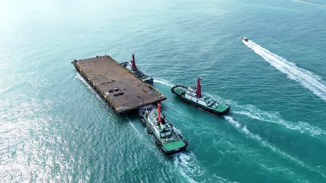 Tugboats towing a large empty flat deck barge through the blue ocean. Heavy marine logistics, offshore construction transport, and commercial maritime shipping operations.
