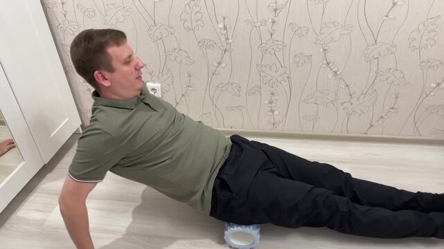 White man performing glute release on foam roller while seated, hands behind for support and legs extended, textured roller under hips for massage and core engagement in casual home interior