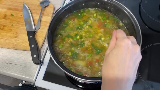 stirring vegetable soup in pot over stove, visible hand using spoon, diced carrots and peas, fresh herbs in simmering broth, wooden cutting board with knives at side, steam rising, spoon tasting