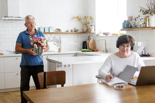 Senior husband giving flowers to working wife on laptop in living room. Domestic life and remote work. Happy Mothers day. Mature business woman receiving bouquet on holiday