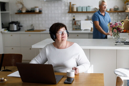 Portrait of senior woman working on laptop at wooden table in living room on background of modern kitchen and husband cooking. Mature business woman freelancer having work at home