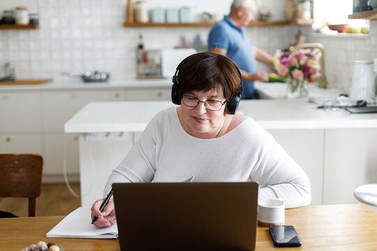 Portrait of senior woman working on laptop at wooden table in living room on background of modern kitchen and husband cooking. Mature business woman freelancer having work at home
