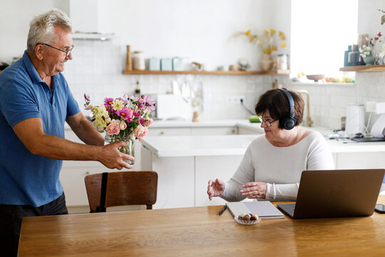 Senior husband giving flowers to working wife on laptop in living room. Domestic life and remote work. Happy Mothers day. Mature business woman receiving bouquet on holiday
