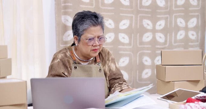 Senior asian entrepreneur leans toward laptop, reviews accounting paperwork, checks online order list, compares invoice with receipt, monitors parcel box stack, manages shipping schedule for delivery.