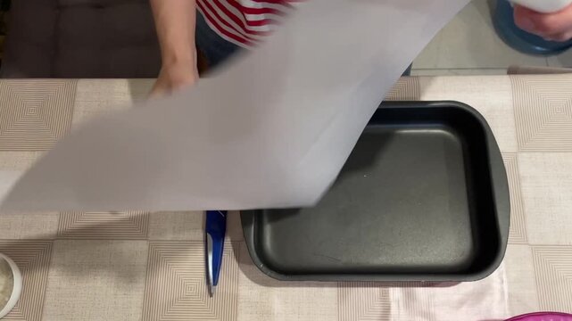 hands lining baking tray with parchment. roll of paper and scissors on patterned tablecloth, home baker smoothing corners into nonstick pan, stepbystep neat preparation for cookies or bars, calm