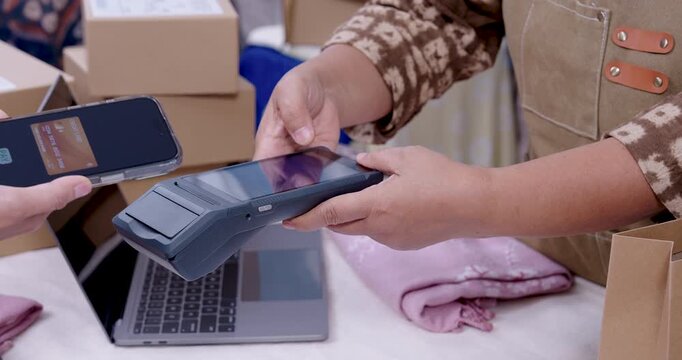 Senior asian woman entrepreneur processes cashless tap to pay on POS terminal, retired woman small business checks invoice on laptop, prepares handmade shibori fabric parcel for shipping delivery.