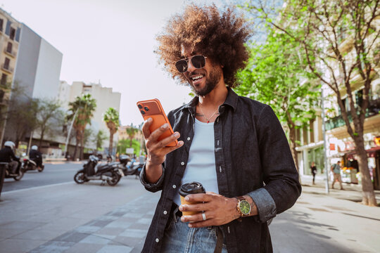 Young latin american man with afro hair, smiling while checking his mobile phone and holding coffee in an urban setting.