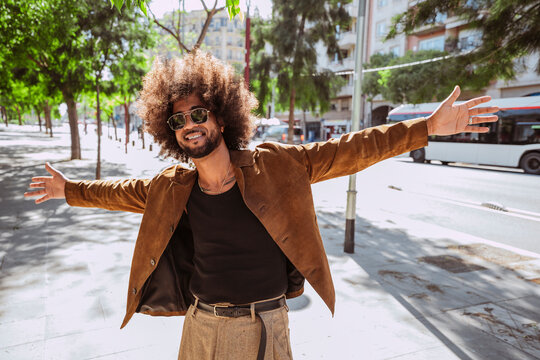 Happy latin american man with afro hair, with outstretched arms standing on a sunny urban sidewalk.
