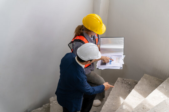 Professional engineers wearing hardhats holding document folder to discuss and inspect staircase construction progress inside residential building site from high angle view