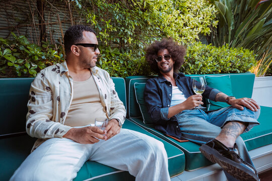 Two latin american male friends relaxing and talking, sharing drinks on a green outdoor couch at terrace surrounded by plants.
