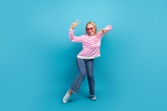 Joyful young woman in pink striped sweater and sunglasses flashing peace signs on bright blue background