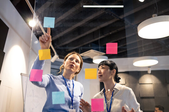 Businesswomen collaborating on glass wall with sticky notes, organizing ideas during corporate brainstorming session