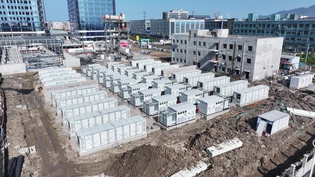 Rows of gray electrical power equipment units arranged neatly on a construction site with city buildings in the background