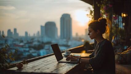Obraz premium Young woman working on laptop at cafe with city skyline sunset view.