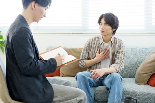 カウンセリング　ソファに座って話をする若い日本人男性と話を聞きながらメモをする若い日本人医師