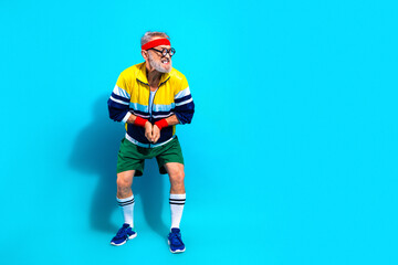 Obraz premium Active senior man in retro sportswear doing a playful exercise pose with headband and wristbands against a bright blue background