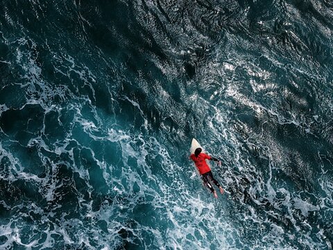 Aerial view of a surfer paddling through deep blue ocean water and white sea foam at Praia do Areal de Santa Barbara in Ribeira Grande, Acores, Portugal.