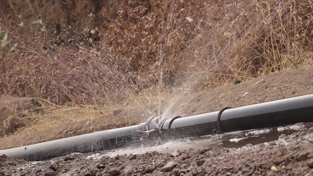 Water spraying from a leaking high-pressure black pipeline connection. Broken water main pipe bursting and leaking outdoors on soil. High-pressure water leak at a plastic pipe joint connection