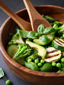 A close-up shot shows a vibrant green salad in a rustic wooden bowl. The salad contains fresh ingredients like broccoli, peas, avocado, and cucumber, garnished with basil leaves and served with wooden