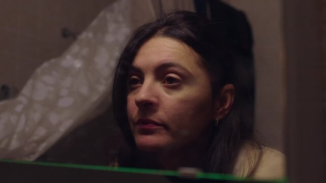 Middle aged woman in 40s staring at reflection in bathroom mirror, pensive mood while reflecting on aging changes and midlife identity at home
